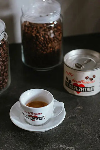 Histórica taza de café blanca Filicori Zecchini de los años 40-1950 - histórico bote de vacío Filicori Zecchini de los años 40-1950 - tarros de cristal llenos de granos de café tostado al fondo.